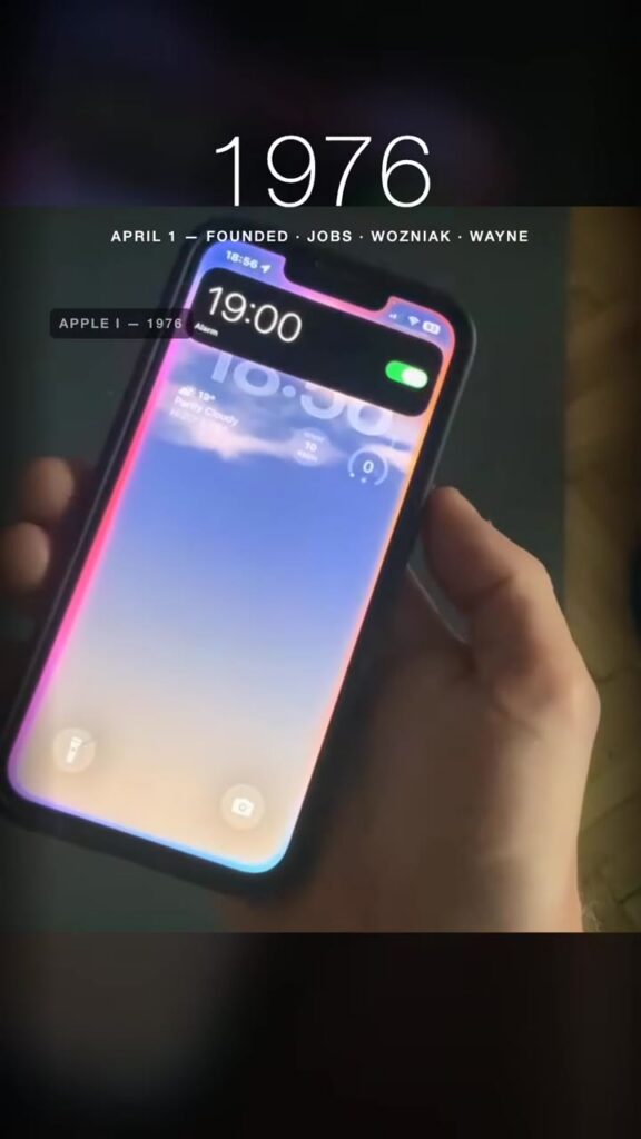 A hand holding an iPhone showing an alarm notification for 19:00, under a caption reading 1976, April 1, founded, Jobs, Wozniak, Wayne.